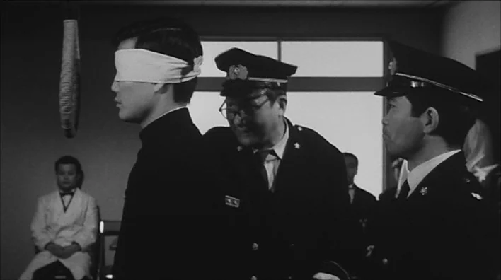 Death By Hanging (1968) movie still: medium — A stark indoor space with a large window and plain light-colored walls. A young blindfolded man stands…