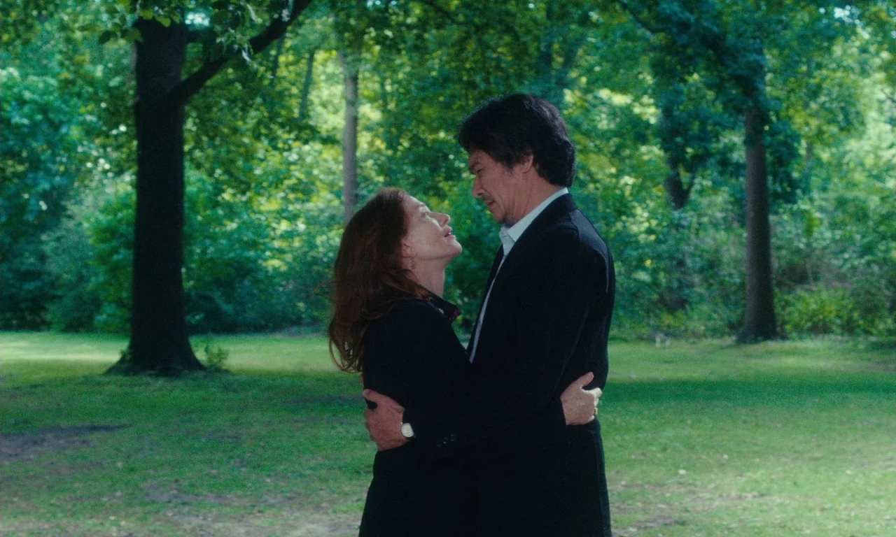 Sidonie in Japan (2023) movie still: medium — A man and woman embrace in a grassy park surrounded by mature deciduous trees. The pair stands in the…