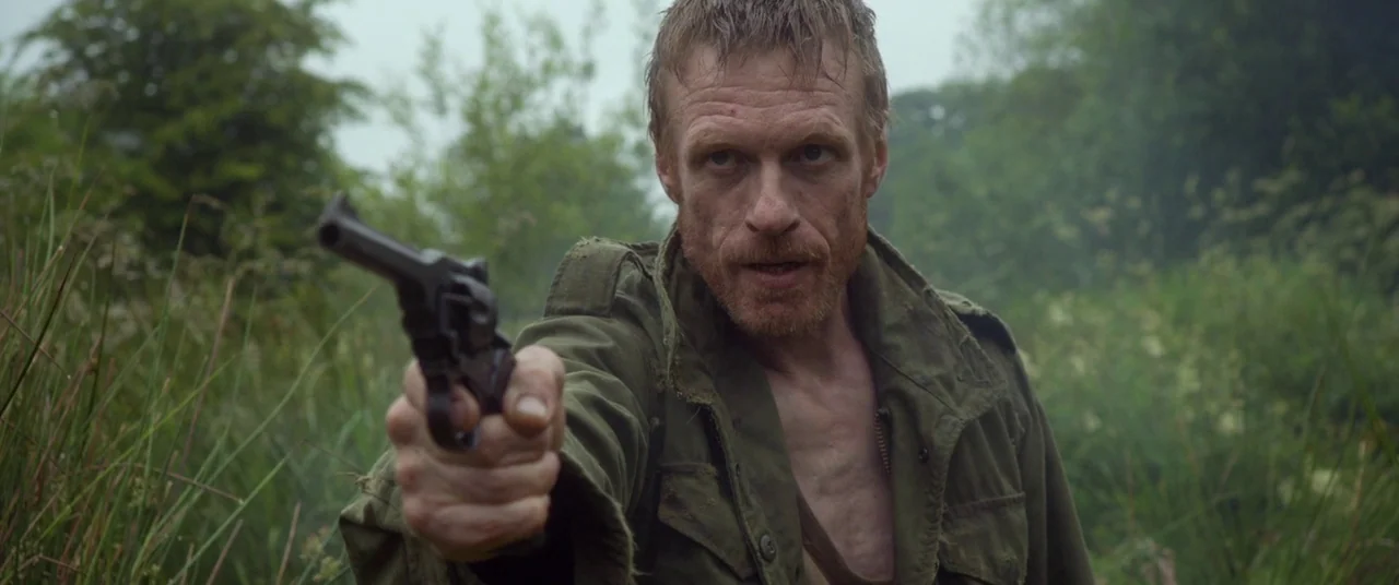 The Survivalist (2015) movie still: medium — An overgrown field provides the background for a man in military attire. The subject stands in the tall…