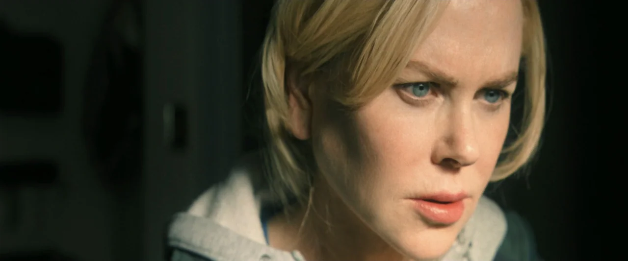 Holland (2025) movie still: closeup — A dimly lit room provides a dark backdrop for a close-up shot. A blonde woman with light eyes focuses her…
