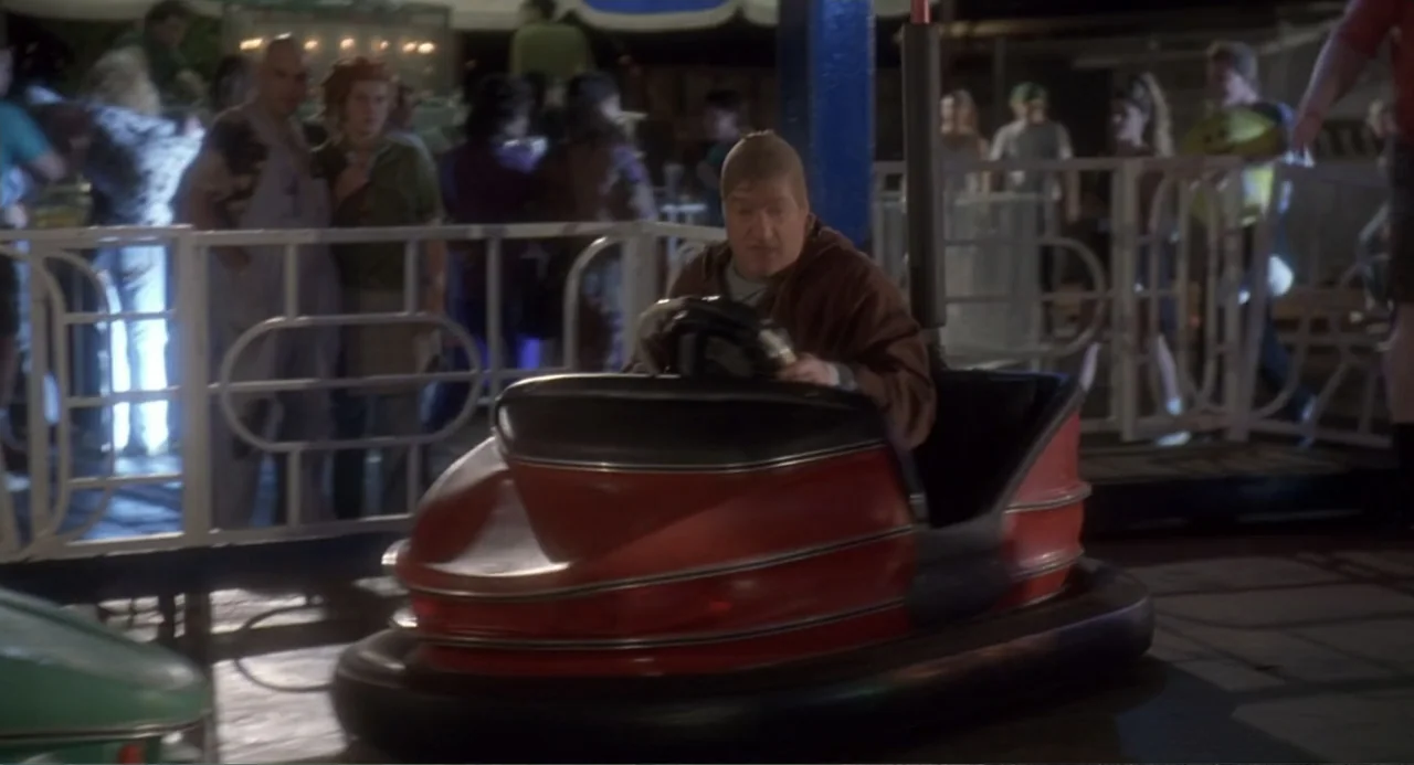 Fatal Instinct (1993) movie still: medium — A night time carnival arena with a metallic white railing bordering the bumper car track. A man wearing…