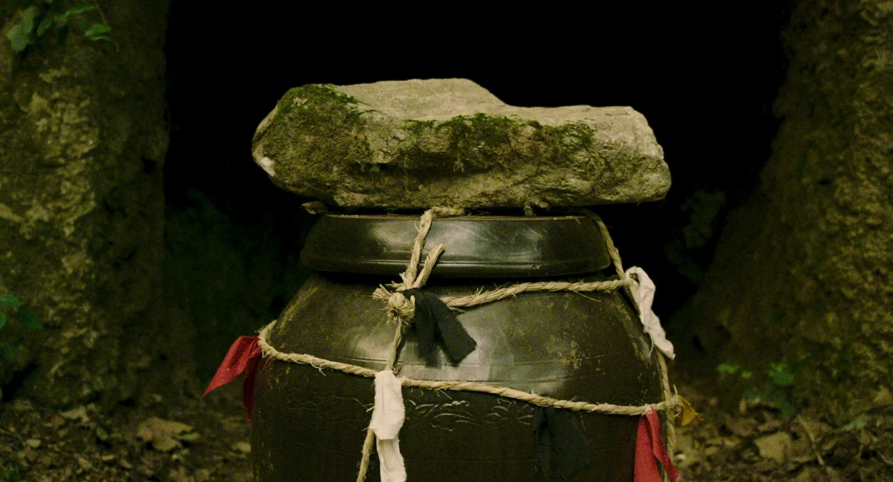 The Mimic (2017) movie still: closeup — A dark outdoor space features a traditional Korean pottery jar wrapped in rope. The jar lid is topped with a…