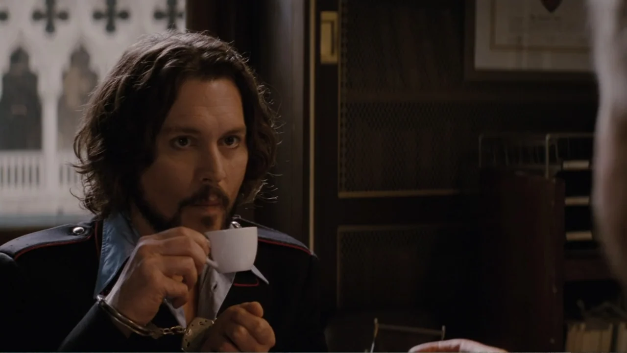 The Tourist (2010) movie still: medium — A man sits at a desk in an office with a view outside. He holds a white espresso cup with handcuffed wrists.