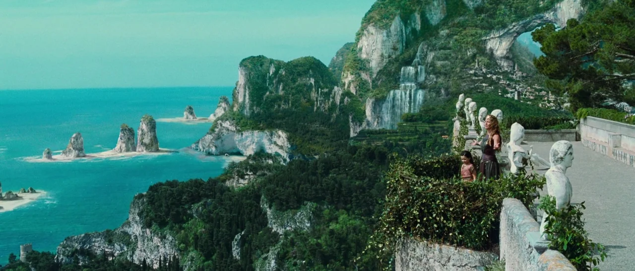 Wonder Woman (2017) movie still: wide — A stone terrace overlooks a vast coastal landscape with towering cliffs and turquoise water. A woman and a…