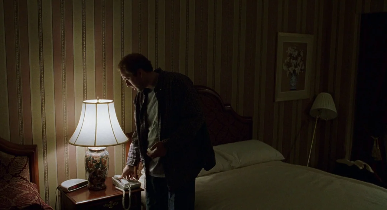 Adaptation (2002) movie still: medium — A dimly lit hotel room features striped wallpaper and framed art above the bed. The subject stands by the…