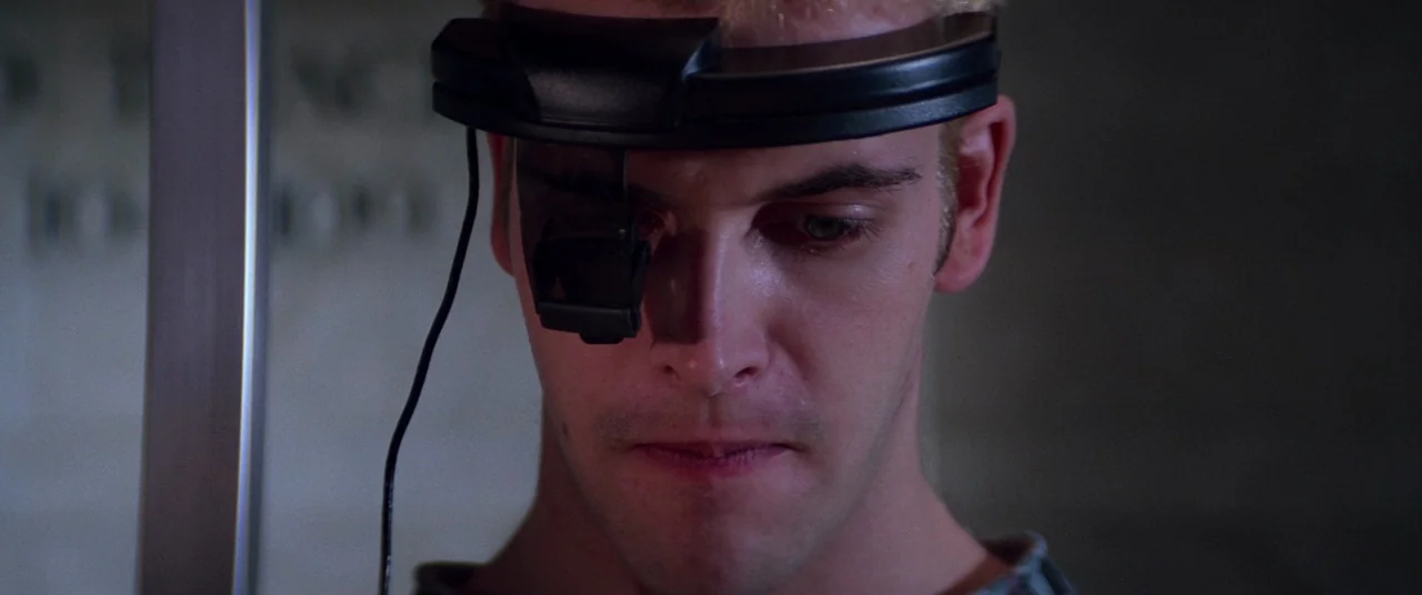 Hackers (1995) movie still: closeup — A man wears a specialized black head-mounted visor device against a grey wall background. He is centered in…