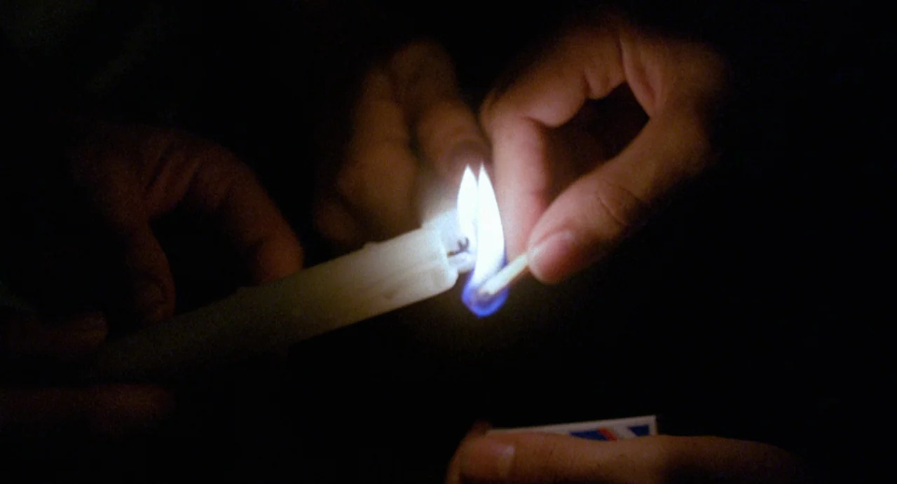 StageFright (1987) movie still: closeup — A close-up view shows two hands working in near total darkness. A wooden match held by one hand lights the…