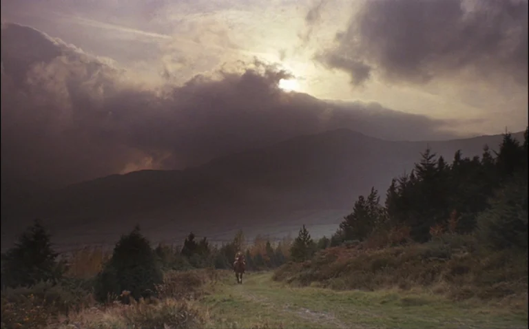 Barry Lyndon (1975) movie still: wide — A desolate mountain landscape features a dirt path bordered by small evergreen trees. A lone rider on…