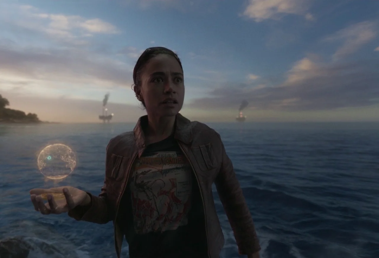 Eternals (2021) movie still: medium — A rocky shoreline at dusk overlooks a deep blue sea with distant industrial oil platforms. A young woman holds…