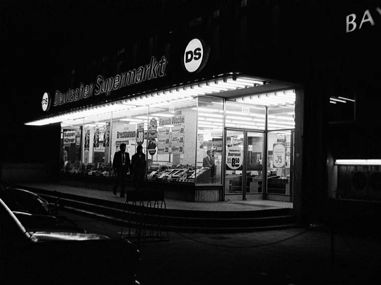 Gods Of The Plague (1970) movie still: wide — A nighttime exterior of a brightly lit German grocery store storefront. The structure features…
