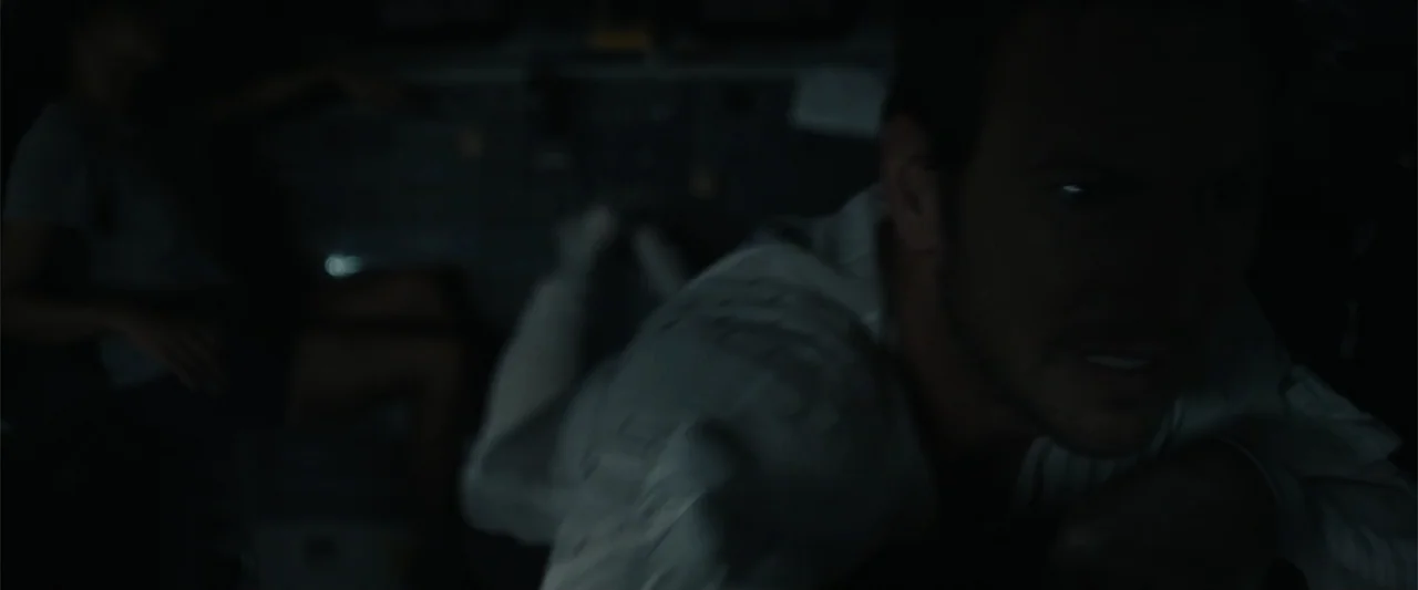 Moonfall (2022) movie still: closeup — A dark, cramped cockpit illuminated only by the faint glow of monitors. A man in a light shirt leans toward…