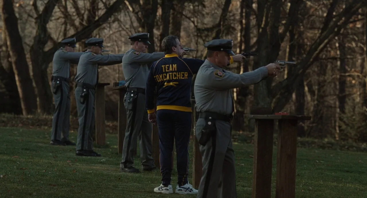 Foxcatcher (2014) movie still: fullbody — A group of men stand at wooden stations in a grassy wooded area. The men hold revolvers directed toward…