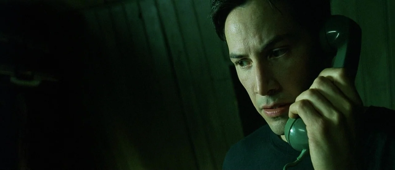The Matrix (1999) movie still: closeup — A man stands in a dark environment with vertical wall panelling visible in background. He holds a vintage…
