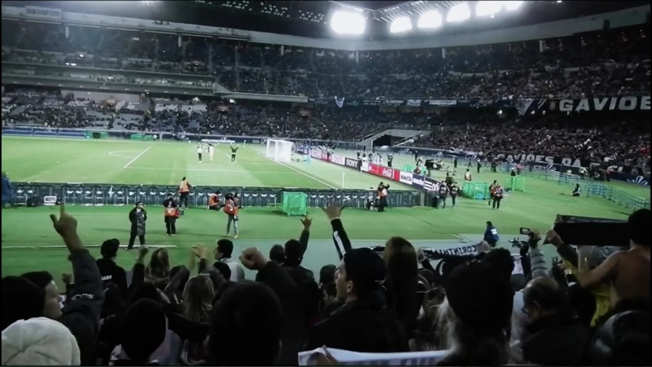 Vai Corinthians (2023) movie still: wide — A large modern soccer stadium is filled with spectators watching a game at night. The crowd in the…