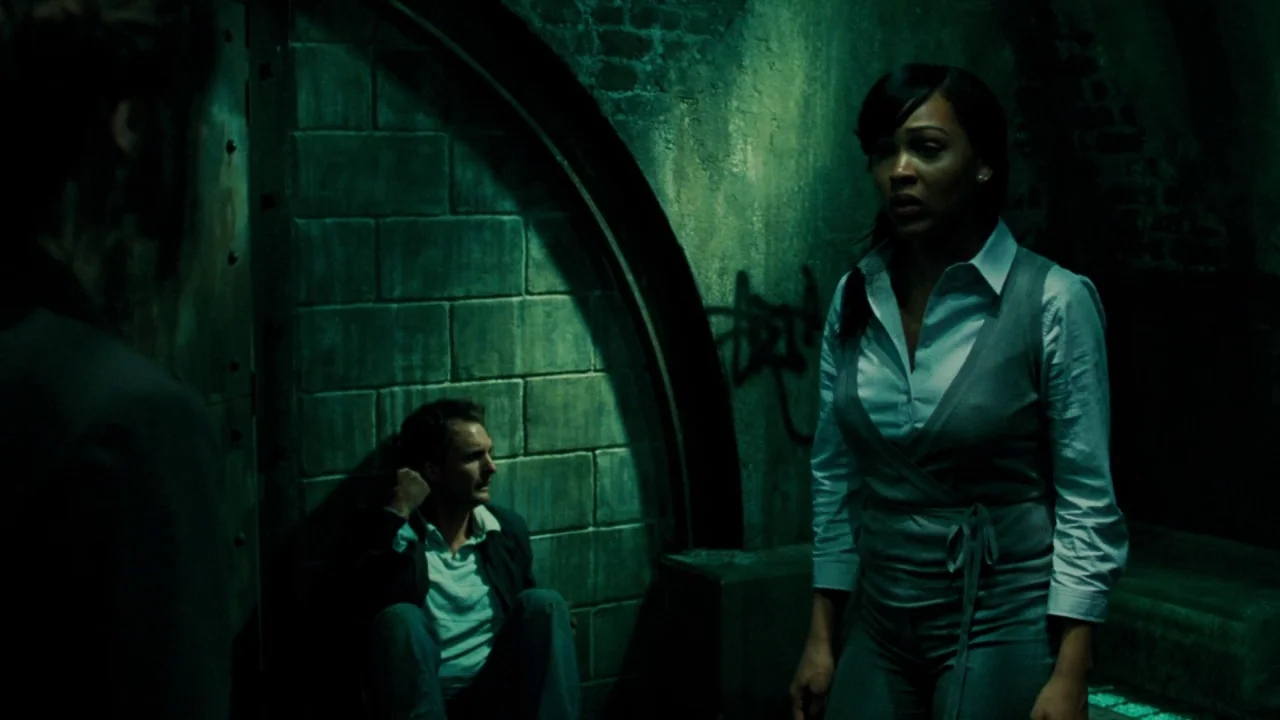 Saw V (2008) movie still: medium — A dark subterranean chamber features damp stone walls with faded graffiti markings. A woman stands in the…