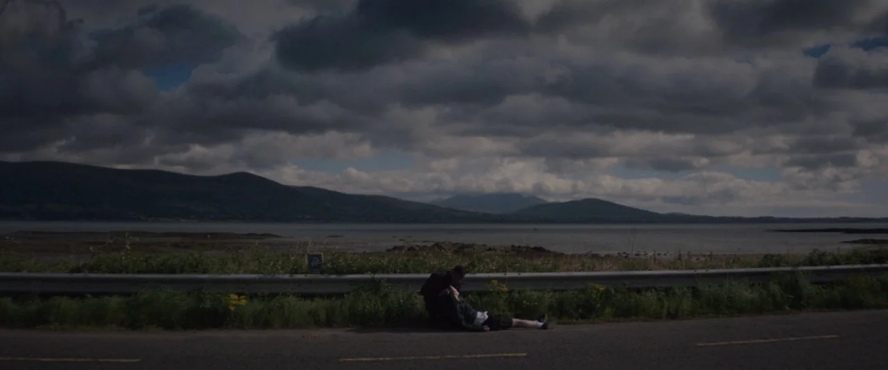 Kissing Candice (2017) movie still: wide — A dark asphalt road separates grassy dunes from a large body of water. One person kneels on the verge…