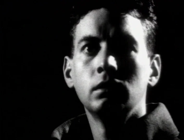 Archangel (1990) movie still: closeup — A dark, unidentifiable space provides no environmental detail behind the human subject. A young male face…
