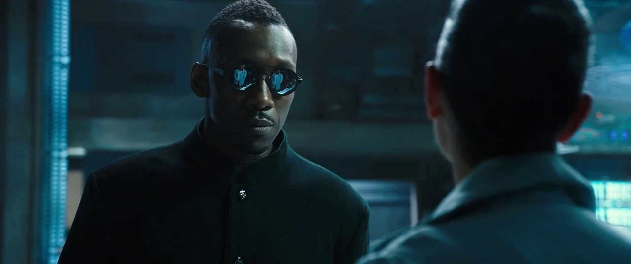 Alita: Battle Angel movie still: medium — A dimly lit room features cool blue displays and technical equipment in the shadows. A man with sunglasses…