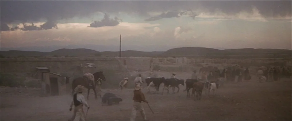 The Wild Bunch (1969) movie still: wide — An expansive, dry, dust-filled field at the edge of a rural settlement under clouds. Several men on foot…