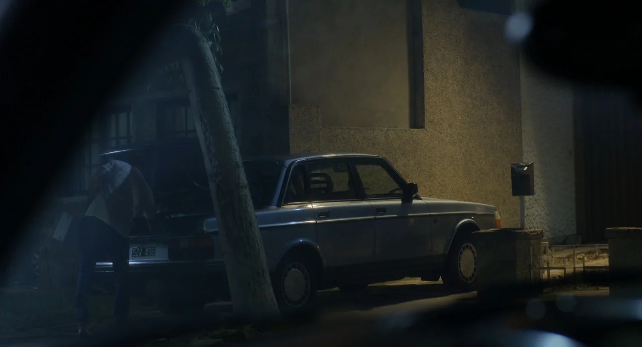 Little White Lies (2010) movie still: medium — A silver sedan is parked at night beside a textured stone building wall. A man stands behind the car…