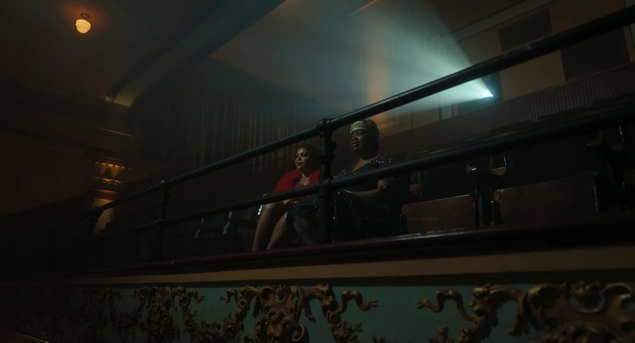 The Color Purple (2023) movie still: wide — An ornate, dimly lit movie theater balcony features gold-leaf moulding along the railing. Two women are…