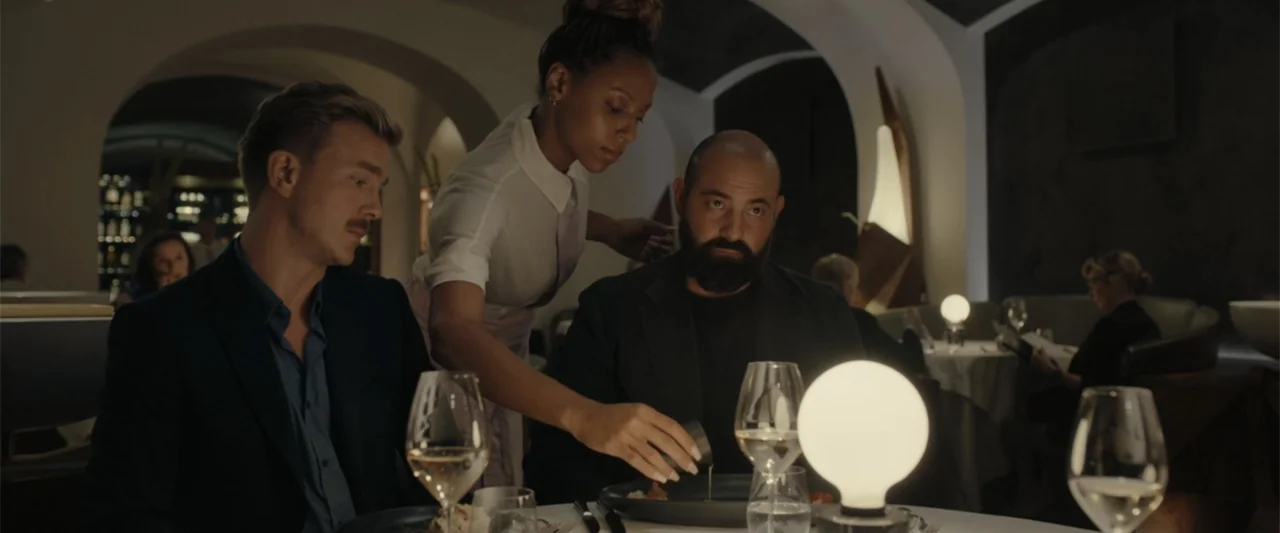 Peacock (2010) movie still: medium — An upscale dining space features rounded white arched walls and dark ambient lighting. A server pours sauce…