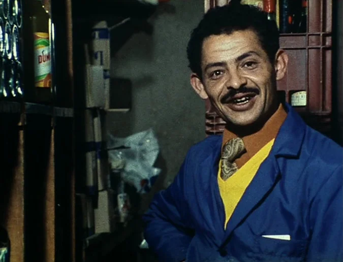 Daguerréotypes (1976) movie still: medium — A man stands inside a small, cluttered retail space filled with shelves and crates. He wears a blue work…