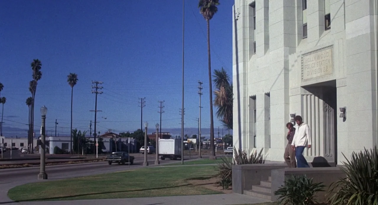 Annie Hall (1977) movie still: wide — A light-colored stone police station building stands beside a street with palm trees. Two men descend the…