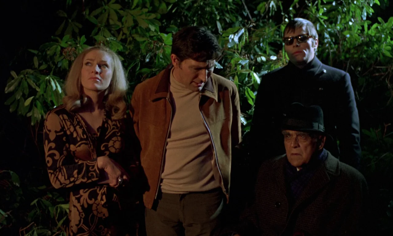 Curse of the Crimson Altar (1968) movie still: medium — Four people stand outdoors at night in front of dense, dark green leafy shrubs. The group…
