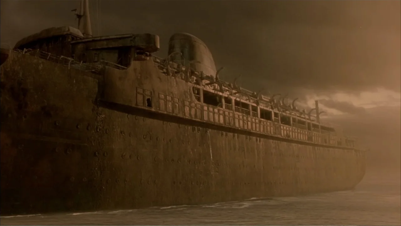 Ghost Ship (2002) movie still: establishing — A massive rusted ocean liner is grounded in shallow water at dusk. The ship looms large against a…