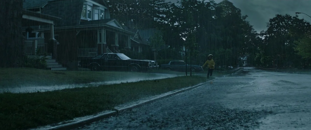 It (2017) movie still: wide — A quiet residential suburban street with houses and parked cars sits under heavy rain. A young child in a bright…