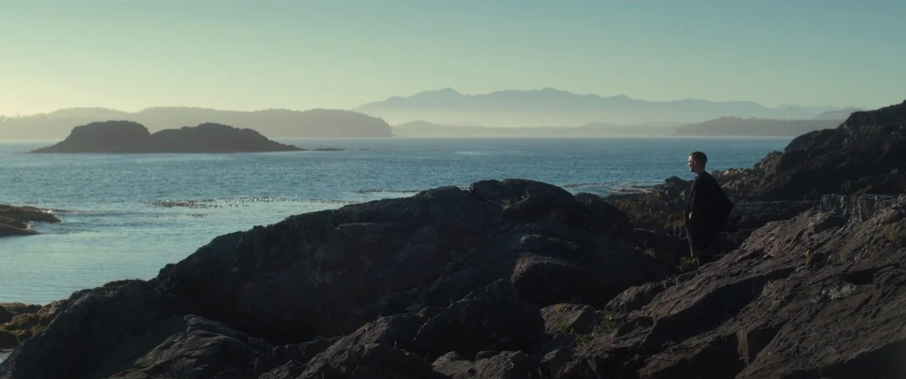 Come To Daddy (2019) movie still: wide — A rugged rocky coastline overlooks a vast ocean with distant mountains in the haze. A lone man in a dark…