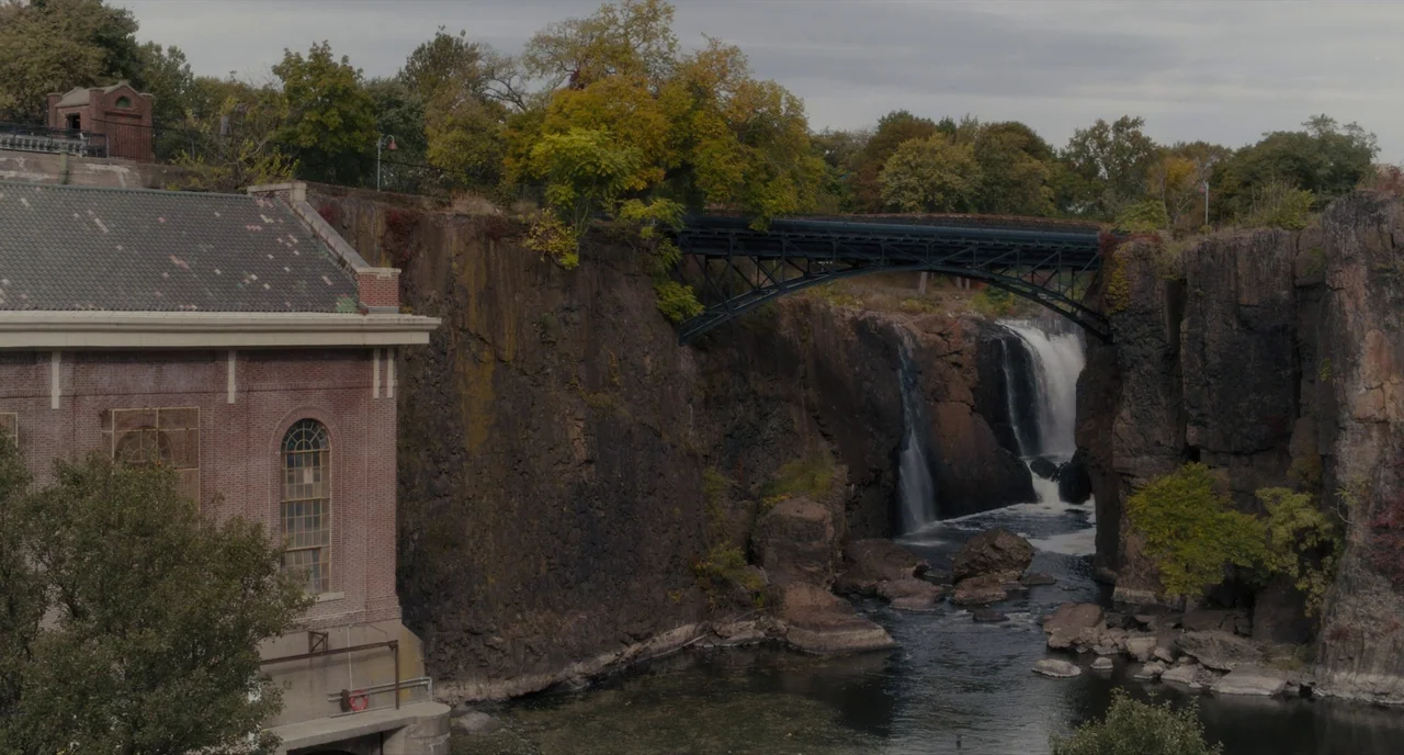 Paterson (2016) movie still: establishing — A brick industrial building stands beside a large waterfall cascading down jagged dark cliffs. A black…