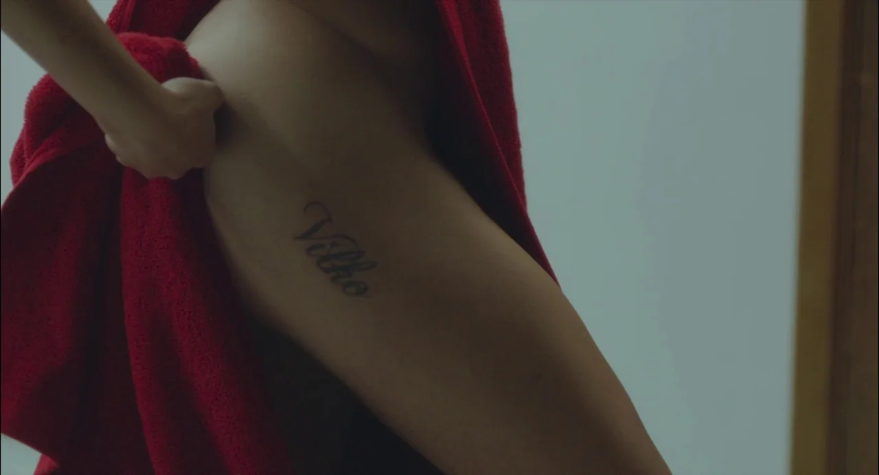 Abuse of Weakness (2013) movie still: closeup — A close view shows a woman's thigh with a visible script tattoo. She grips a bright red towel…