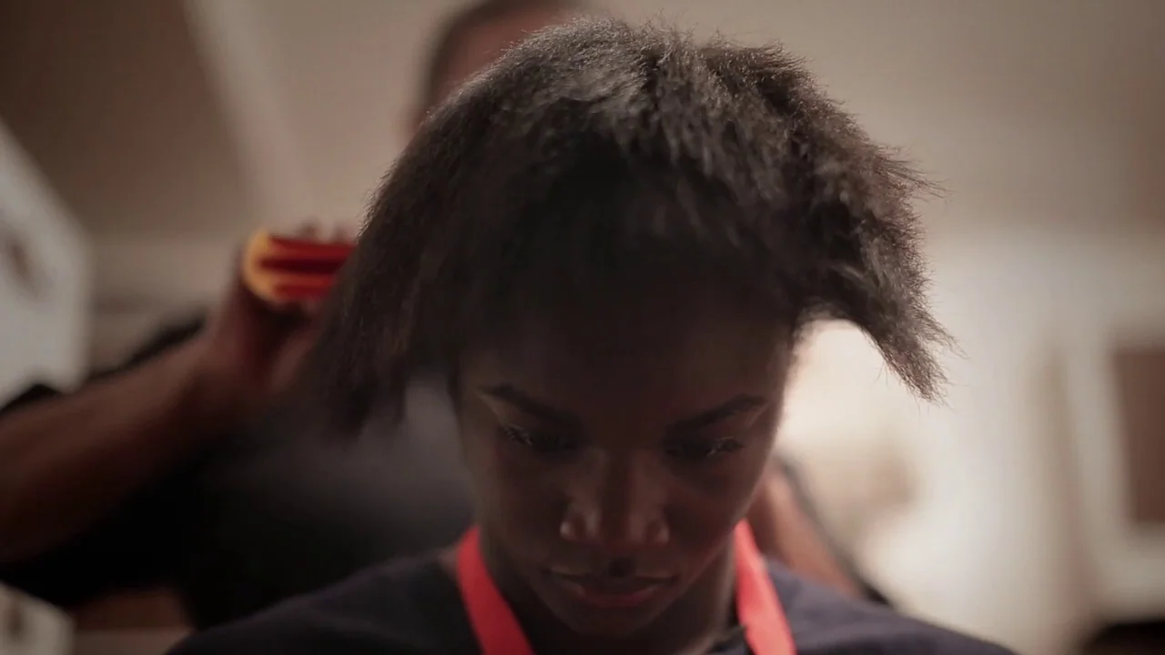 T-Rex (2015) movie still: closeup — A close-up view shows a person getting their hair styled inside a home. The focus remains on the young woman…