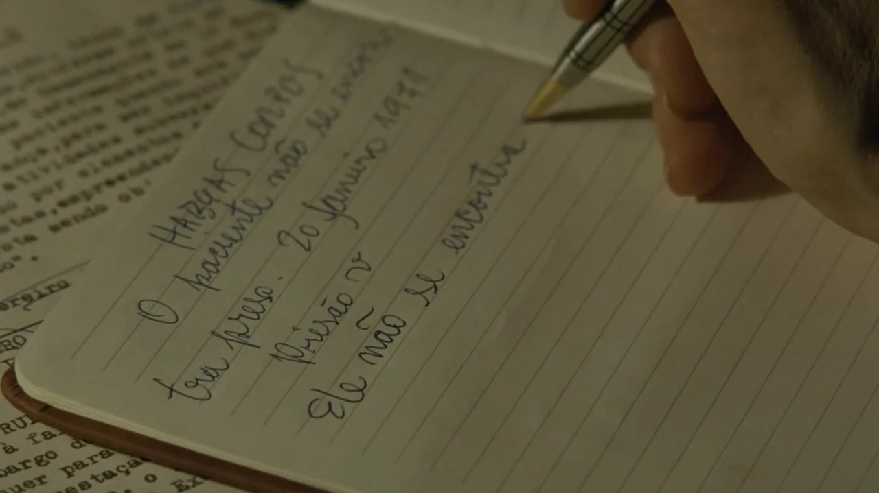 I'm Still Here (2024) movie still: closeup — A close view of a hand writing with a metal pen on a lined notebook page. The notebook sits atop older…