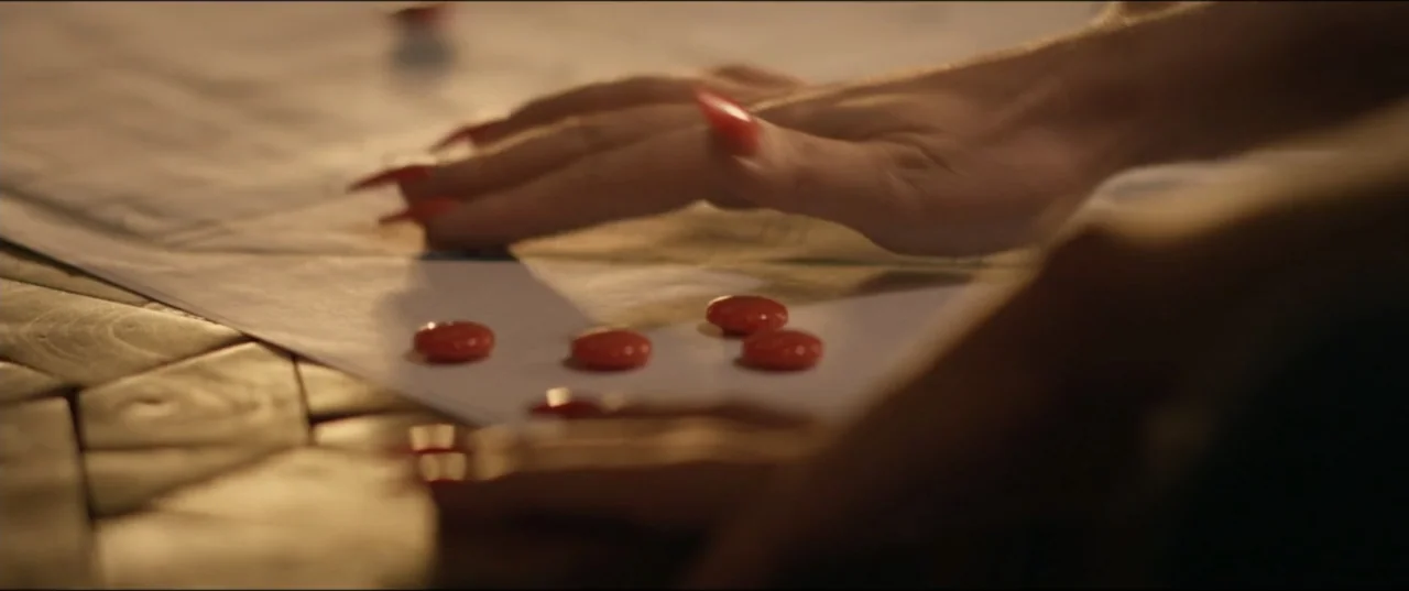 Risqué (2025) movie still: closeup — A close view of hands resting on a table surface covered with paper documents. Small round red tokens are…