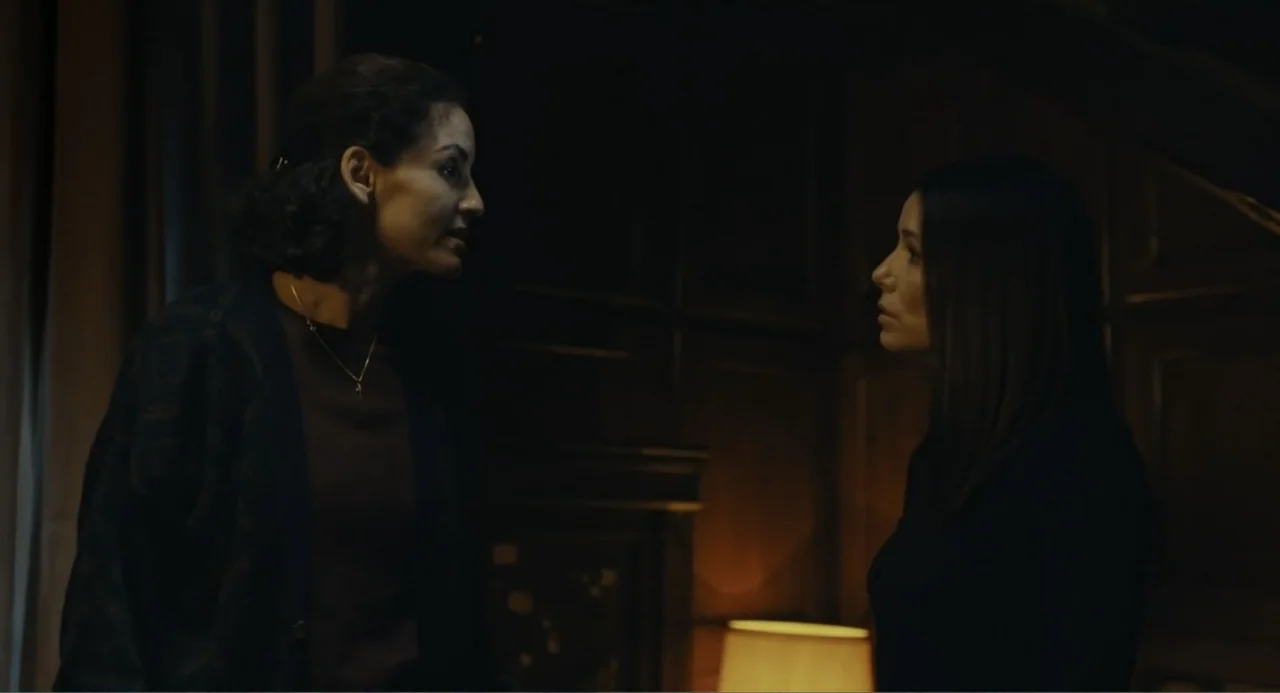 Elles (2018) movie still: medium — A dimly lit room features wood-paneled walls and a single warm practical lamp. Two women stand in profile facing…