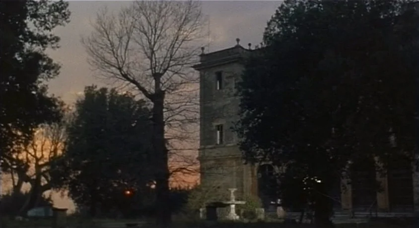 Kill, Baby Kill (1966) movie still: establishing — A stone villa with a prominent tower sits amidst dark, dense trees at dusk. The sky behind the…
