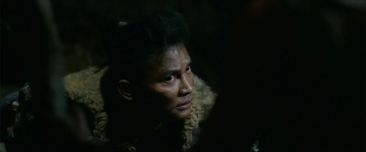Monster Hunter (2020) movie still: closeup — A dark cave interior contains a man illuminated by a nearby firelight source. The man looks forward…