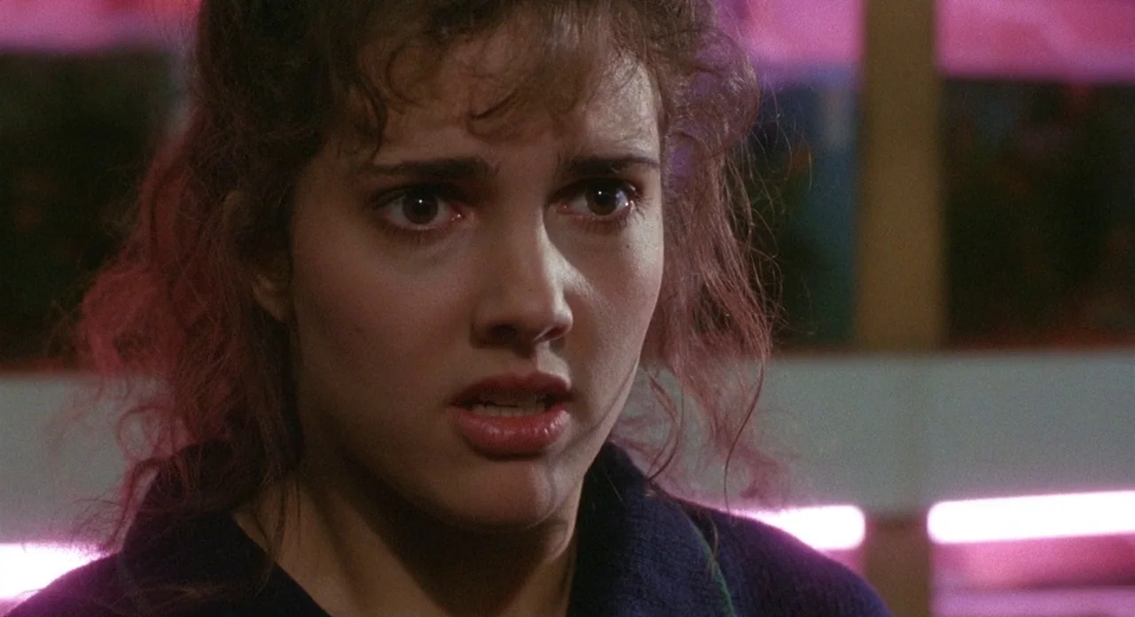 Hellraiser (1987) movie still: closeup — A woman stands in a dim store environment with blurred shelving units behind her. She looks forward with…