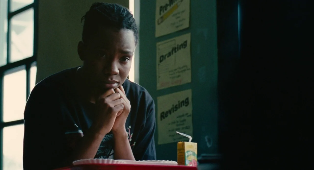 Pariah (2011) movie still: medium — A student sits at a desk in a classroom with posters pinned to a wall. The boy rests his hands together under…