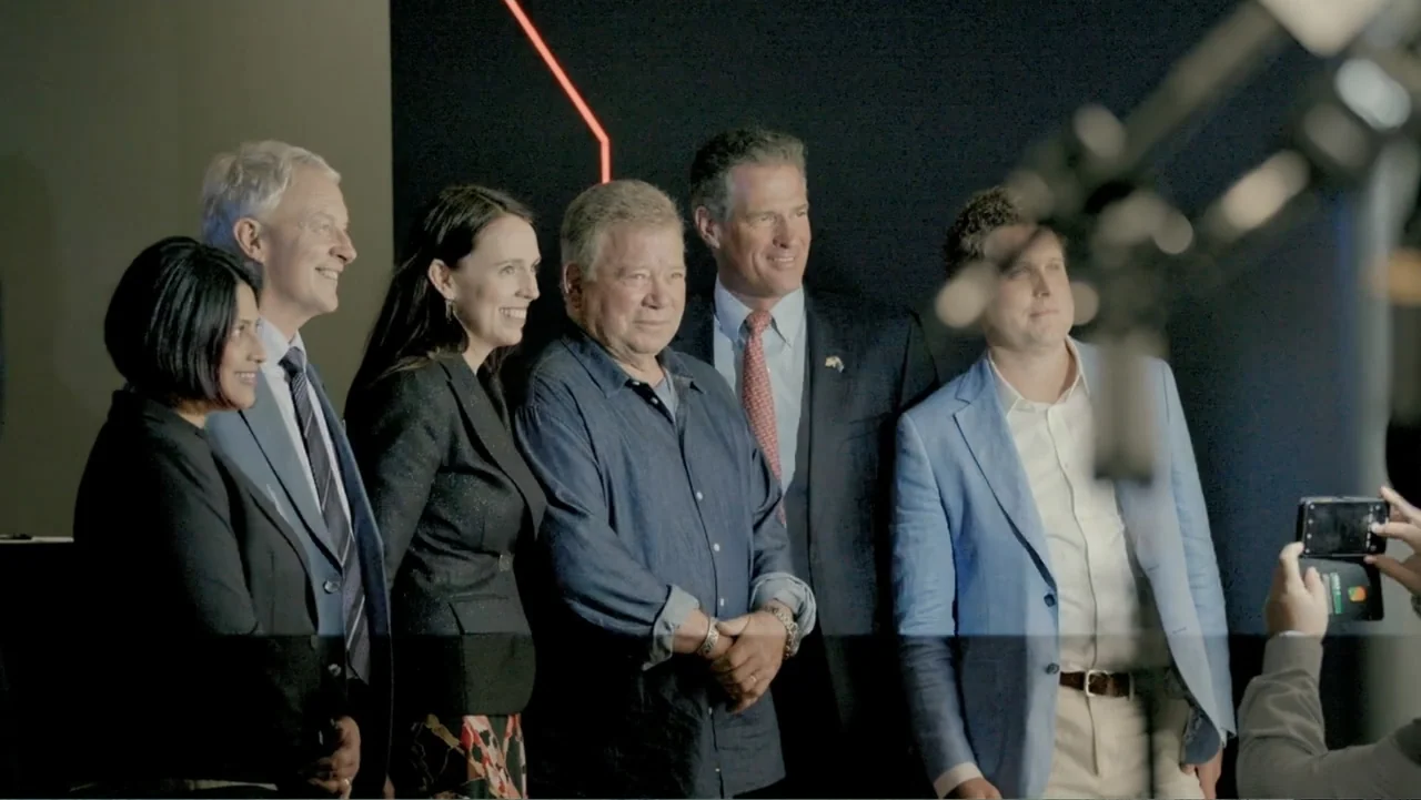 Wild Wild Space (2024) movie still: medium — A group of six people stand side-by-side against a dark wall for a photo. A person in the foreground…