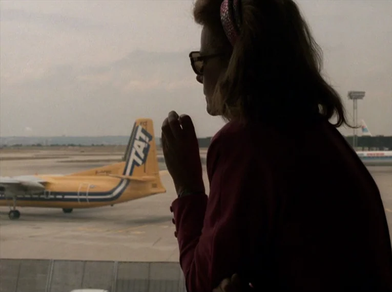 Keep Your Right Up (1987) movie still: medium — An interior airport terminal window looks out onto a concrete tarmac with airplanes. A woman in a…