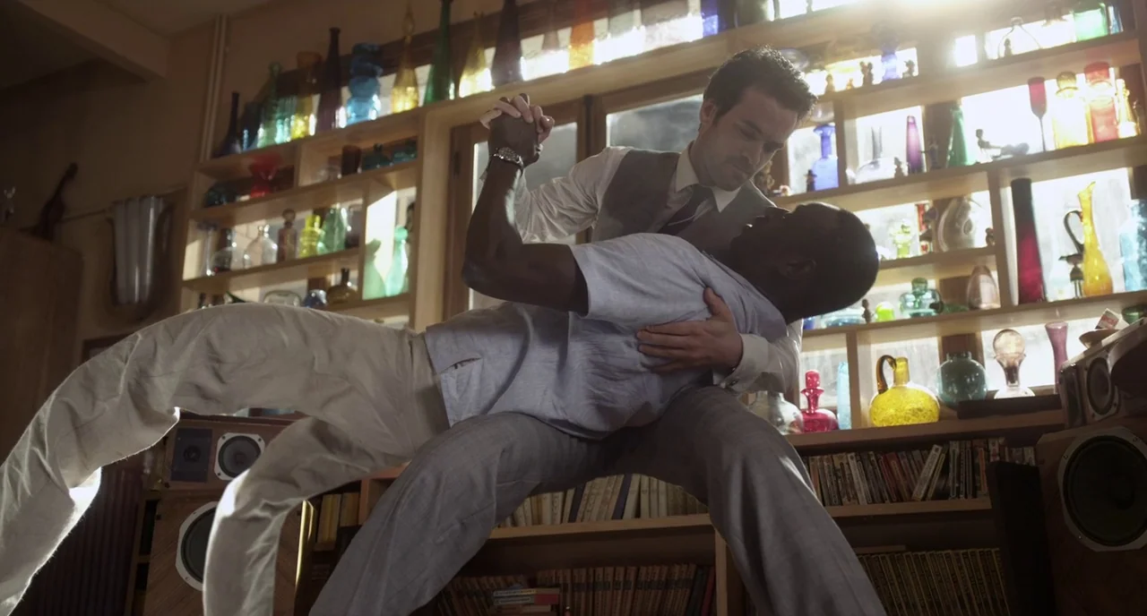 Mood Indigo (2013) movie still: fullbody — A room features extensive wooden shelving units filled with various colorful glass vases. Two men perform…