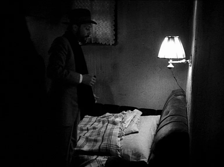 My 20th Century (1989) movie still: medium — A dark, spartan room features a single bed illuminated by a wall sconce. A man in a suit and hat stands…