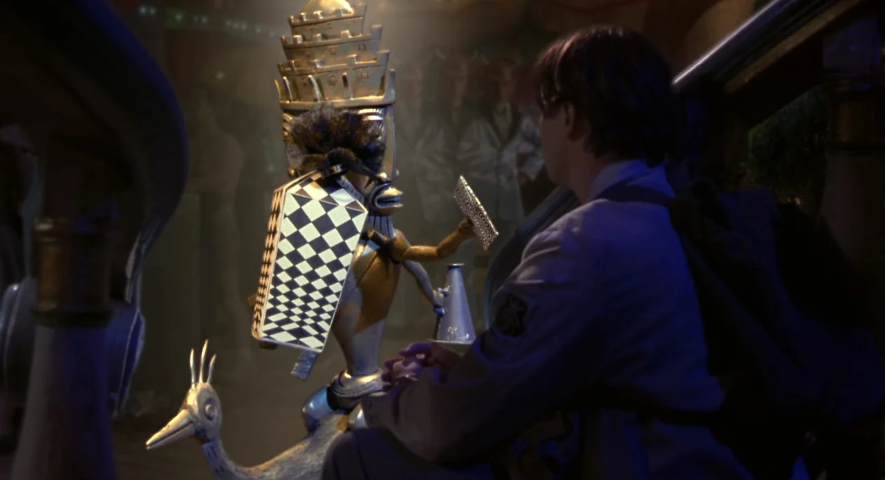 Monkeybone (2001) movie still: medium — A dimly lit room features a man sitting beside a bizarre golden automaton. The automaton holds a megaphone…