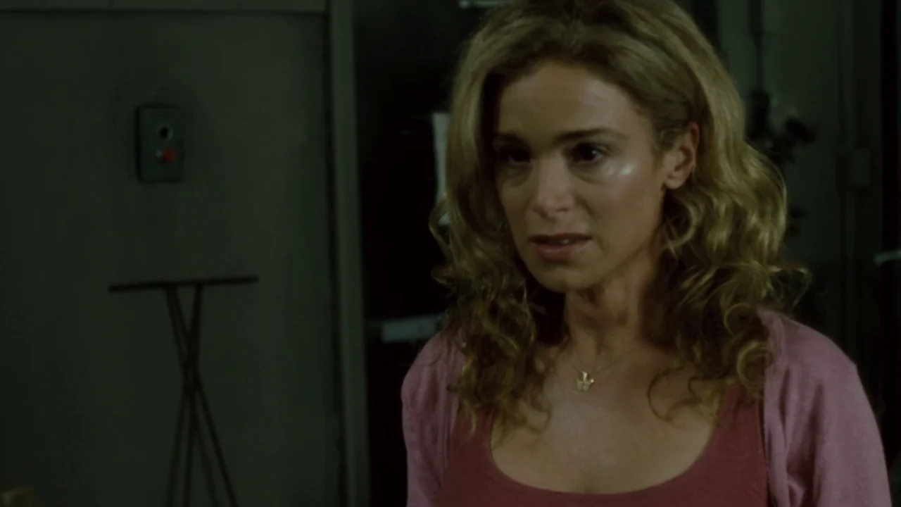 Saw IV movie still: medium — A dark industrial workspace with metal walls and a small control panel switch. A woman with blonde hair wearing a pink…