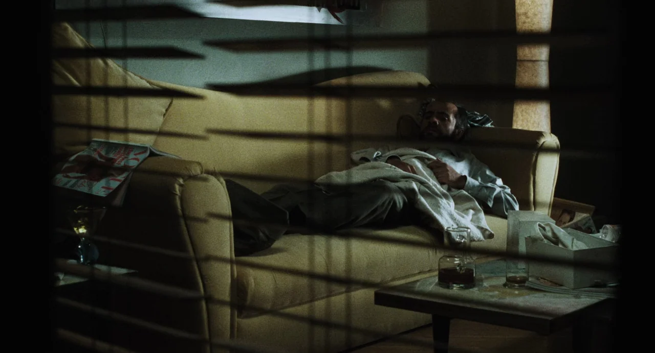 Amores Perros (2000) movie still: medium — A man lies on a yellow sofa viewed through horizontal Venetian blinds. A coffee table with a glass…