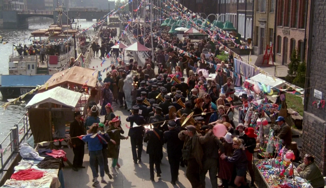 The Rainbow Thief (1990) movie still: establishing — A bustling outdoor market occupies the paved path alongside a river with boats docked. A dense…
